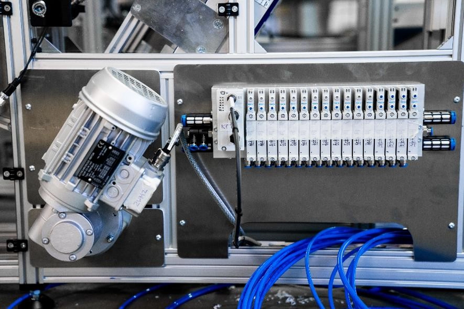 WI Automtisierungstechnik GmbH - Industrielle Automatisierung - Industrieanlage mit einem silbernen Elektromotor und einem auf einer dunkelgrauen Platte montierten Getriebe. Rechts neben dem Motor befindet sich ein pneumatischer Ventilblock mit mehreren Ventilen und angeschlossenen blauen Luftschläuchen.
