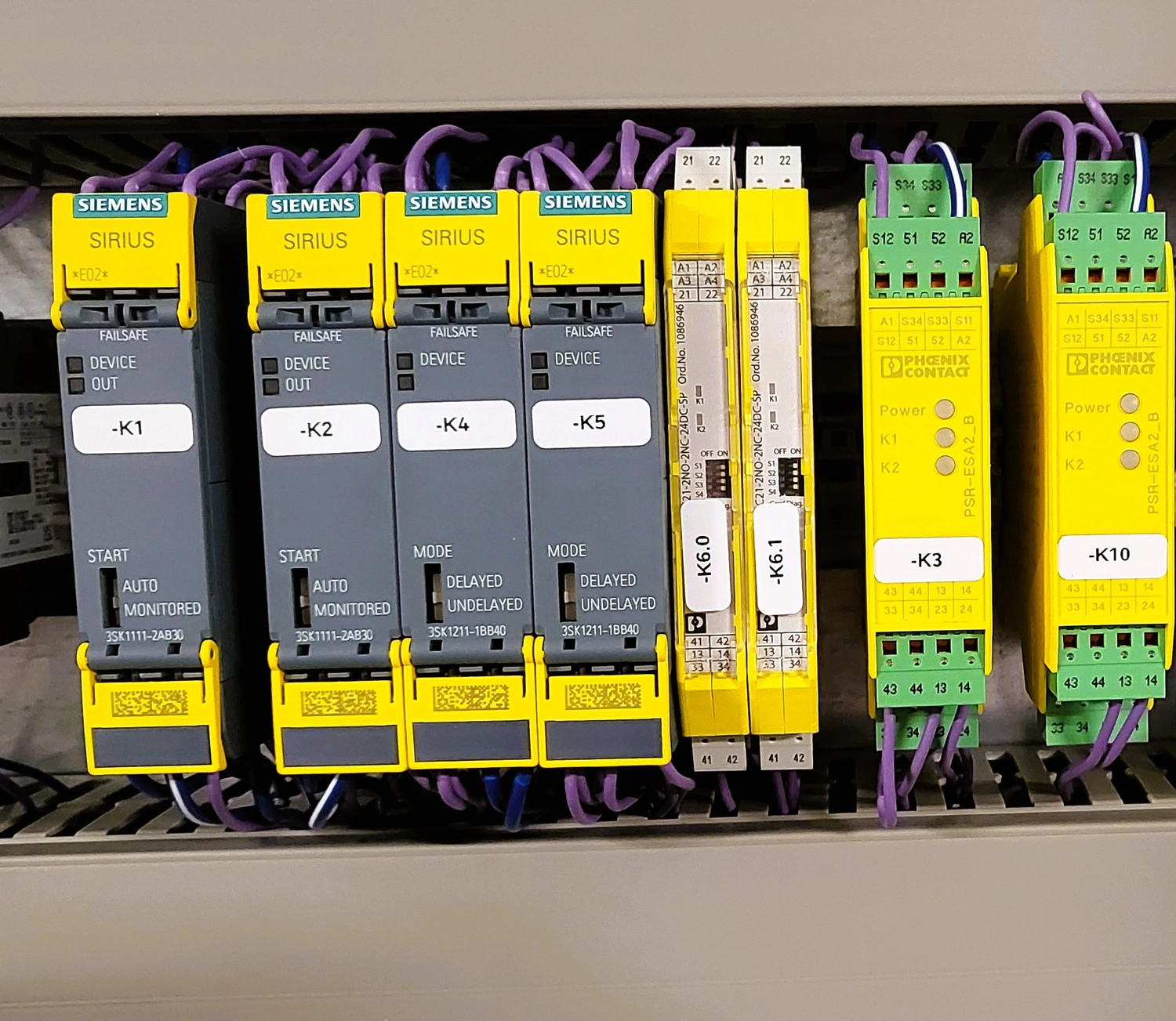 WI Automtisierungstechnik GmbH - Industrielle Automatisierung - Nahaufnahme mehrerer gelber Siemens SIRIUS Sicherheitsrelais und Motorstarter, die auf einer DIN-Schiene montiert sind, mit Beschriftungen wie "-K1", "-K2", "-K4", "-K5" und "-K6.1".